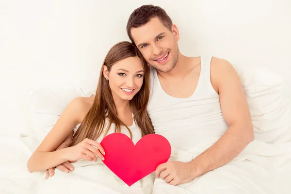 Ευτυχισμένο ζευγάρι στην αγάπη ξαπλωμένη στο κρεβάτι και κρατώντας κόκκινο χαρτί καρδιά — Φωτογραφία Αρχείου