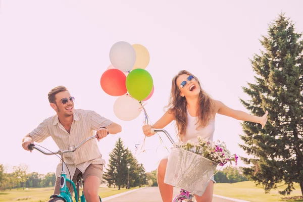Alegres amantes felizes andando de bicicleta com balões e flores — Fotografia de Stock