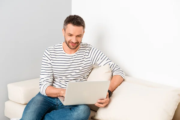 Hombre guapo sentado en el sofá y leyendo noticias en el portátil — Foto de Stock