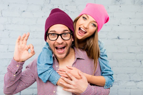 Cute girl huging her boyfriend and boy in glasses gesturing "OK" — Stock Photo, Image