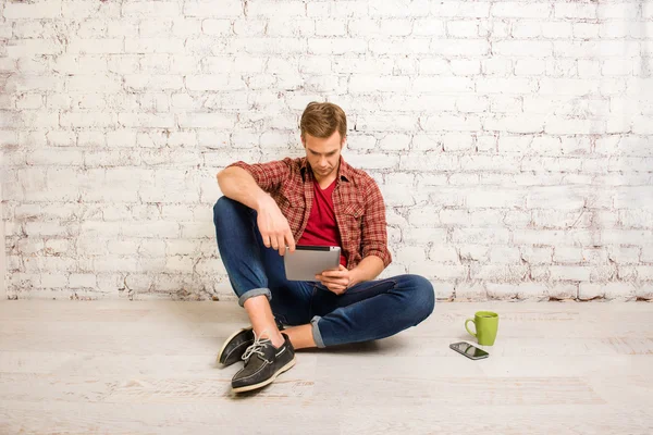 Joven sentado en el suelo y leyendo noticias en internet en la mesa — Foto de Stock