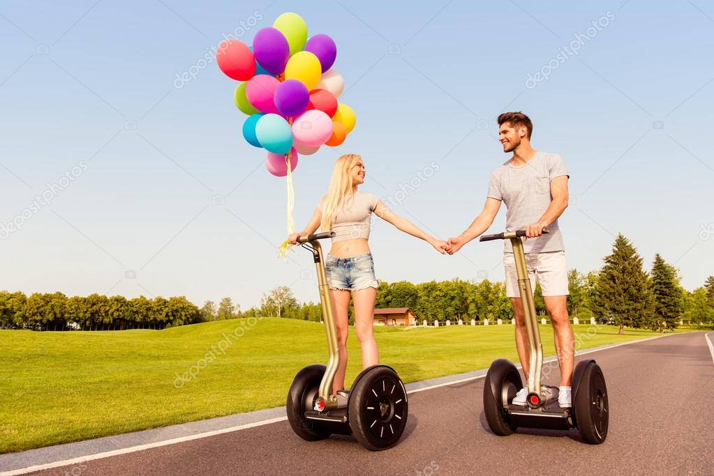 Young cute couple with balloons holding hands on segways in par