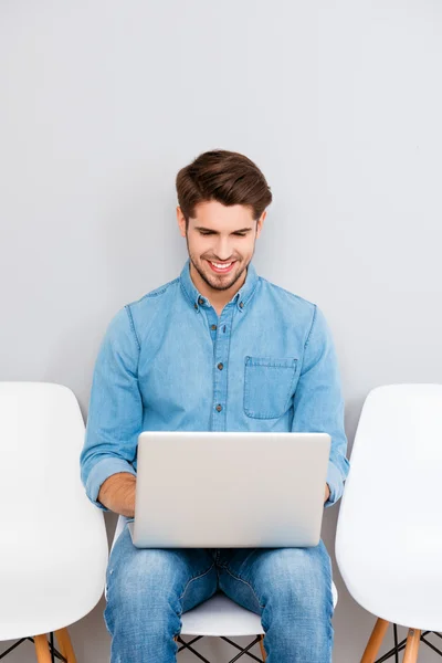 Χαρούμενος νεαρός άνδρας κάθεται σε καρέκλα και να εργάζονται με φορητό υπολογιστή — Φωτογραφία Αρχείου