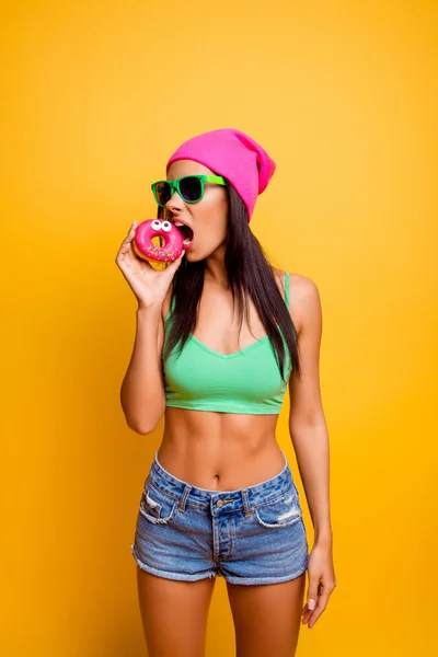 饿了有趣的女孩，在粉色的帽子咬眼睛附近 y 粉色甜甜圈 — 图库照片