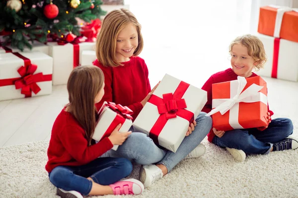 Portret van drie mooie aantrekkelijke mooie vrolijke kinderen zitten op tapijt vloer uitwisselen giftboxen Eve Noel hulst vrolijke dag in licht wit interieur appartement huis binnen — Stockfoto