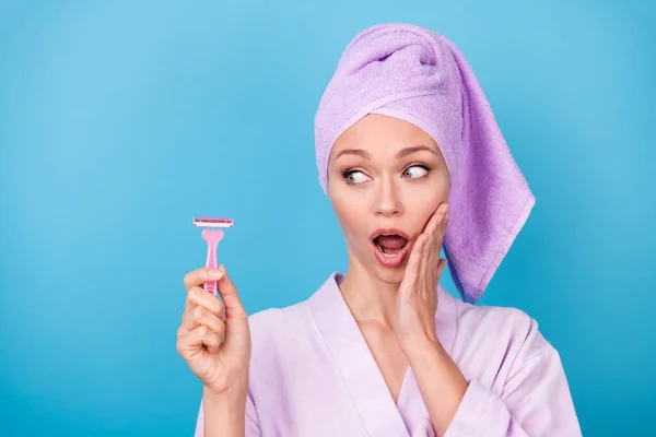 Fénykép a csinos nő néz oldalán tartsa rózsaszín borotva kar arccsont nyitott száj viselni lila törölköző turbán fürdőköpeny elszigetelt kék háttér — Stock Fotó