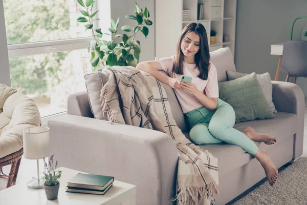 Foto retrato de la mujer sosteniendo el teléfono en dos manos sentado con ambas piernas en el sofá en el interior — Foto de Stock