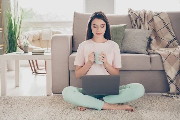 Foto ritratto di donna seduta sul tappeto pavimento in posa di loto con il computer portatile che tiene la tazza in due mani a guardare video al chiuso — Foto Stock