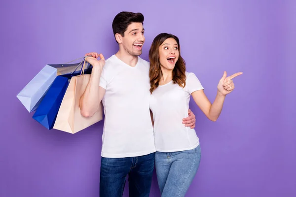 Foto hübsche Dame Kerl schockiert niedrige Preise Paar gute Laune einkaufen gehen halten viele Geschäft Paket Spree direkte Finger leer Raum tragen lässige T-Shirts Jeans isoliert lila Farbe Hintergrund — Stockfoto