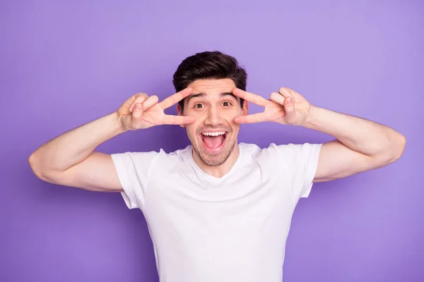 Photo przystojny atrakcyjny facet student zębaty uśmiech zabawny wyraz twarzy pokazując v-signs w pobliżu oczy szalony beztroski osoba nosić casual biały t-shirt odizolowany fioletowy kolor tła — Zdjęcie stockowe