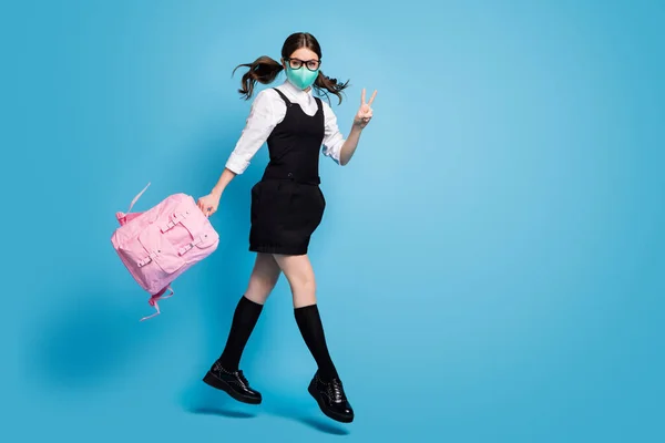 Full size photo of dream high school girl jump up hold rucksack rucksack rucksack make v-sign wear white blouse black dress rock overall long socks medical mask isolated blue color background — Stockfoto