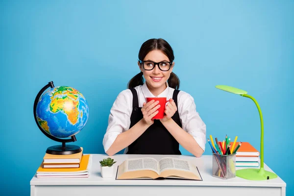 Foto av positiv high school flicka sitta bord håll dryck mugg njuta av akademiska kurser lärande bära vit blus svart övergripande uniform isolerad över blå färg bakgrund — Stockfoto