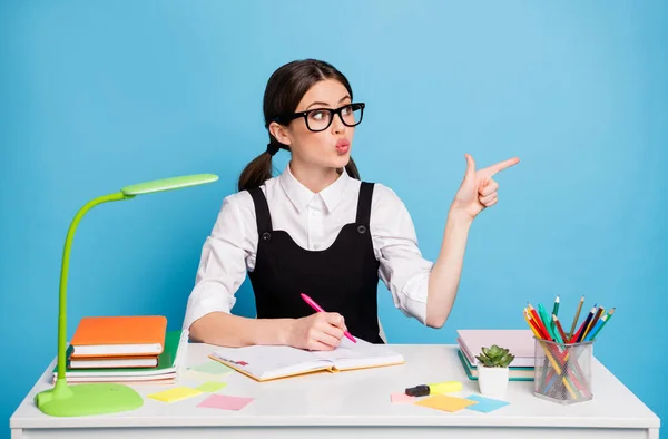 Foto de menina do ensino médio espantado sentar mesa escrever copybook ponto indicador dedo copyspace fazer lábios despojado desgaste branco preto blusa uniforme isolado sobre azul cor de fundo — Fotografia de Stock