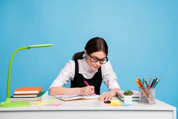 聪明集中的女孩坐在桌子上写抄本高中毕业设计身穿白色黑色时髦制服与蓝色背景完全隔离的照片 — 图库照片