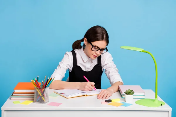 Foto da menina focada do ensino médio sentar mesa escrever copybook tarefa de casa para cursos acadêmicos desgaste branco preto uniforme geral isolado sobre fundo de cor azul — Fotografia de Stock