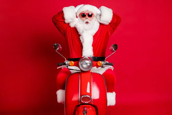 Foto van gepensioneerde opa witte baard handen hoofd geschokt late kerstavond moeten sneller vervoer rit retro bromfiets slijtage x-mas kostuum jas zonneglas kap geïsoleerde rode kleur achtergrond — Stockfoto