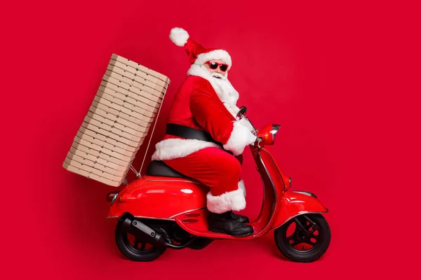 Volledige lengte profiel foto van gepensioneerde opa witte baard rijden pizza dozen ondersteunen sponsoring nieuwjaar evenement dragen santa x-mas kostuum jas zonneglas cap laarzen geïsoleerde rode kleur achtergrond — Stockfoto