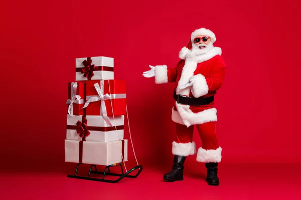 Полная длина фото отставной старик белая борода рекомендуя сани много Giftbox лучший выбор носить Санта-мас костюма пальто солнцезащитное стекло головной убор черные сапоги изолированный красный цвет фона — стоковое фото