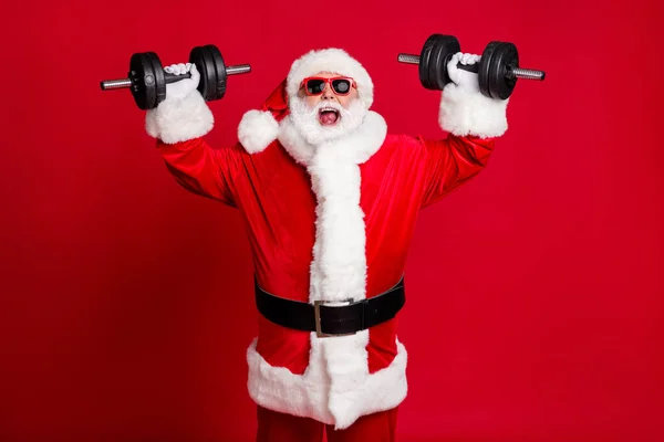 Zdjęcie emeryta stary szalony człowiek biały broda podnieś hantle otwarte usta krzycząc ciężka waga ignorując ból mięśni nosić Santa X-mas kostium czapka okulary przeciwsłoneczne odizolowany czerwony kolor tło — Zdjęcie stockowe