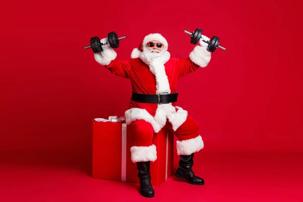 Full length photo of retired old man white beard hold dumbbells sit large box training before trip north pole wear santa costume sunglass headwear leather boot isolated red color background — Stock Photo, Image