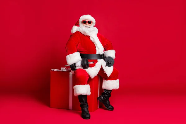 Full length photo of retired old man white beard sit big giftbox hold dumbbells warming up arms wear x-mas santa costume glove coat belt sunglass cap boot isolated red color background — Stock Photo, Image