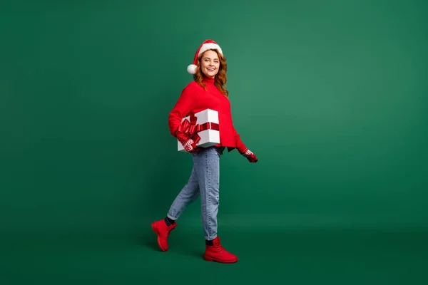 Foto de perfil de comprimento total da senhora go carry giftbox wear red sweater x-mas headwear jeans boots isolated green color background — Fotografia de Stock