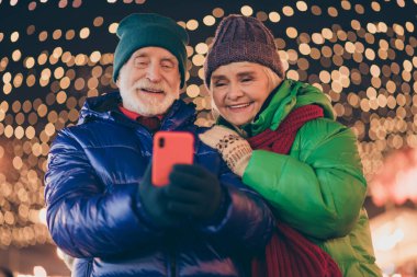 Photo of two people pensioner lovers man woman hold telephone watch funny pictures videos liking give feedback wear coat red scarf headwear x-mas night street illumination fair outdoors outside clipart