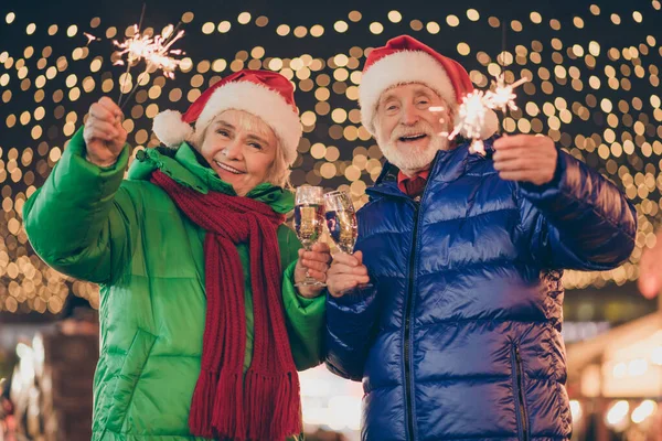 Foto av två personer pensionerade par man kvinna möte hålla tomtebloss vinglas dricka vin vänta chiming klocka midnatt bära rock röd halsduk mössa x-mas kväll gata belysning rättvis utomhus — Stockfoto