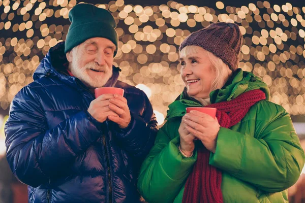 二つの灰色の白い髪の古い人の両親は、カップルは、芳香族飲料マグカップを保持外の夜の照明の下でクリスマススピリットウォークの日付を楽しむ結婚 — ストック写真
