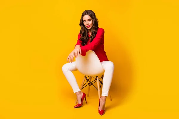 Полная длина фото привлекательной деловой леди сидеть уютный стул между ног серьезно смотреть камера носить красную рубашку белые брюки на высоких каблуках изолированные желтый яркий цвет фона — стоковое фото