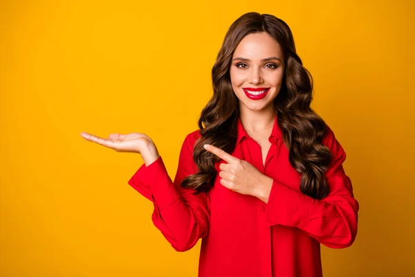 Photo of pretty curly business lady hold open hand empty space sales manager oferta nowość produkt direct finger arm wear red office shirt isolated yellow bright color background — Zdjęcie stockowe