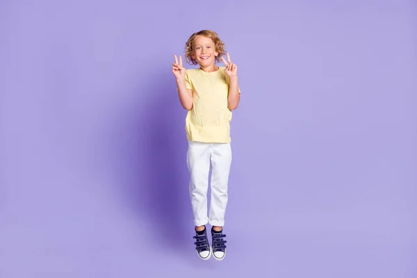 Фотографія позитивного маленького хлопчика, що підстрибує, робить v-знак біло-жовтою футболкою ізольованою на фіолетовому кольоровому фоні — стокове фото