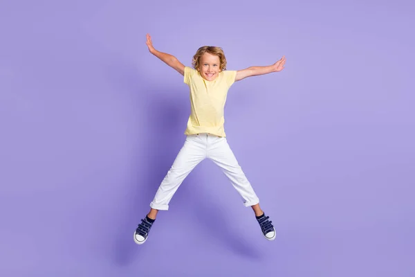 Фотографія веселого хлопчика, який стрибає вгору, тримає руки, носить жовтий білий одяг, ізольований на фіолетовому кольоровому фоні — стокове фото