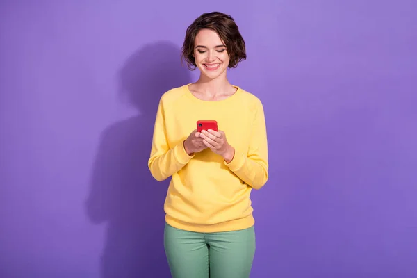 照片上的积极女孩阅读社交媒体的消息智能手机穿套头衫裤子隔离紫色背景 — 图库照片