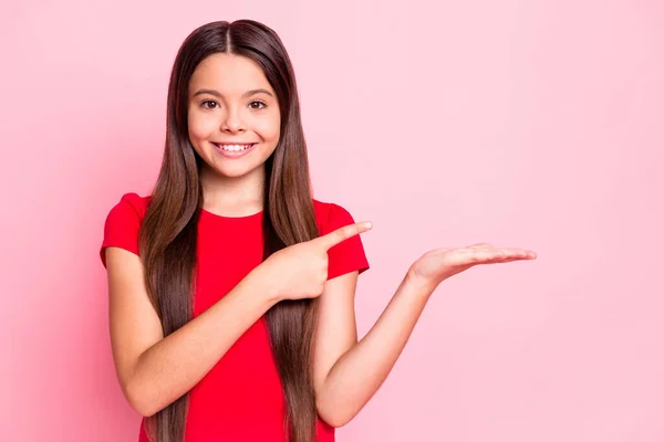 Foto de encantador doce positivo pequena senhora penteado longo braço da palma segurando espaço vazio direto indicar dedo irradiando sorrindo desgaste vermelho t-shirt isolado cor rosa fundo — Fotografia de Stock