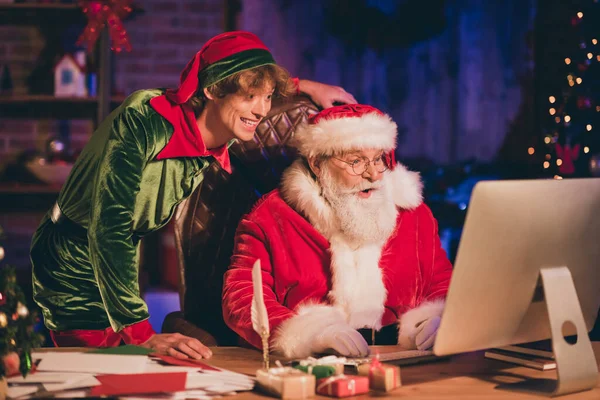 Фотопортрет Санта-Клауса и эльфа, просматривающего интернет на рабочем столе — стоковое фото