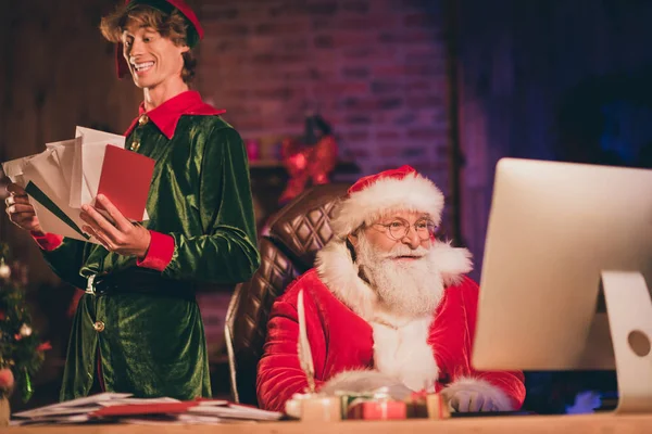 Фотопортрет Санта-Клауса, просматривающего интернет на рабочем столе и эльфа, держащего бумажную почту — стоковое фото