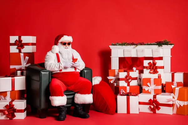 Porträt seiner er schön attraktiv funky Fett übergewichtig Santa mit Gerät Gadget sitzt im Sessel in dekorierten Raum isoliert über hellen lebendigen Glanz lebendigen roten Farbhintergrund — Stockfoto