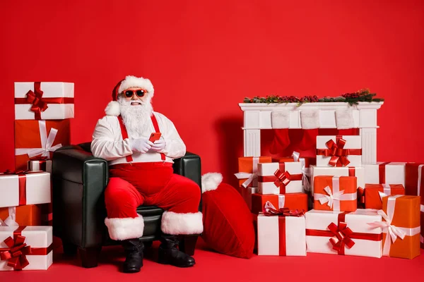 Porträt seiner er schön attraktiv funky Fett fröhlich Weihnachtsmann mit Gadget Blogging Post sitzen im Sessel stapeln Geschenke bestellen isoliert hell lebendigen Glanz leuchtenden roten Hintergrund — Stockfoto