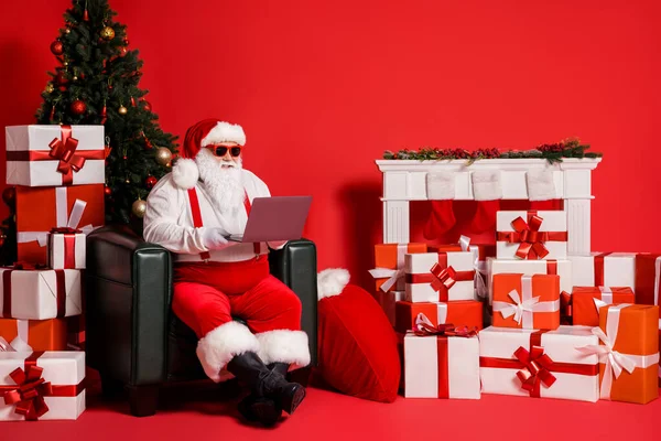 Retrato dele ele agradável atraente gordura funky alegre Pai Natal usando laptop sentado em poltrona leitura lista de desejos e-mail pós entrega do presente isolado brilhante brilho vivo cor vermelha vibrante fundo — Fotografia de Stock