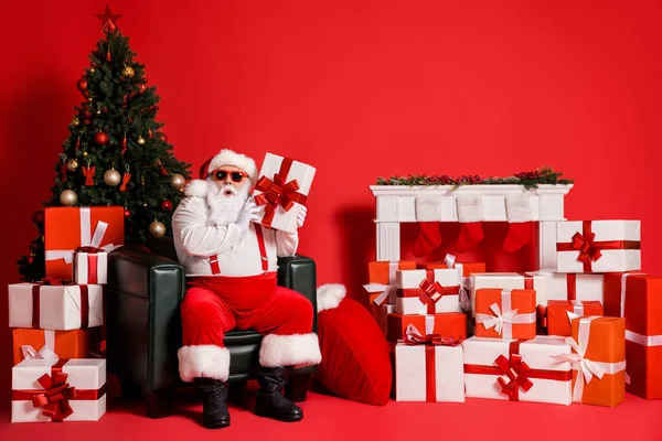 Portret van zijn hij mooi aantrekkelijk funky vet verbaasd benieuwd nieuwsgierig Kerstman zitten in fauteuil vasthouden in handen giftbox geïsoleerde helder levendig glans levendige rode kleur achtergrond — Stockfoto