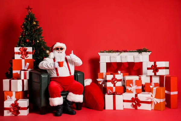 Portret van zijn hij mooie aantrekkelijke funky vet vrolijke Kerstman zitten in fauteuil drinken drank tonen thumbup pauze geïsoleerde heldere levendige glans levendige rode kleur achtergrond — Stockfoto