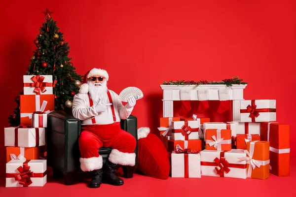 Porträt seiner er schön attraktiv funky fat fröhlich reiche reiche Weihnachtsmann sitzt im Sessel hält in den Händen demonstrieren Bündel Bargeld isoliert hell lebendigen Glanz leuchtenden roten Hintergrund — Stockfoto