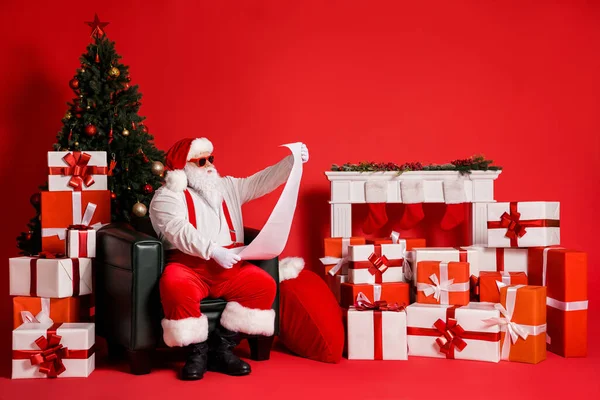 Portret van zijn hij aantrekkelijke funky vet overgewicht Kerstman zitten in fauteuil versierd woonkamer lezen lange papieren brief wishlist geïsoleerde heldere levendige glans levendige rode kleur achtergrond — Stockfoto