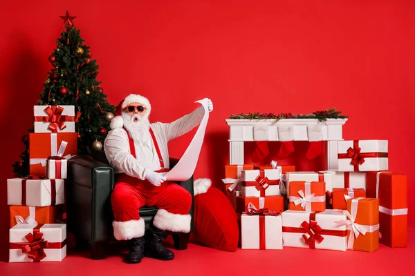 Çekici, şişman, afallamış Noel Baba 'nın portresi oturma odasında oturmuş uzun kağıt mektup hediye kutusu yığınını okuyor parlak, parlak, parlak, kırmızı bir arka plan. — Stok fotoğraf