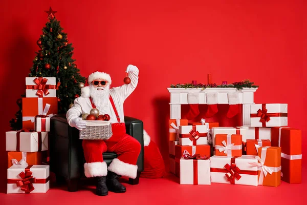 Portret van zijn hij aantrekkelijke funky vet overgewicht vrolijke Kerstman zitten in fauteuil vasthouden in handen ballen feestelijke decor versieren woonkamer geïsoleerde heldere levendige glans levendige rode kleur achtergrond — Stockfoto