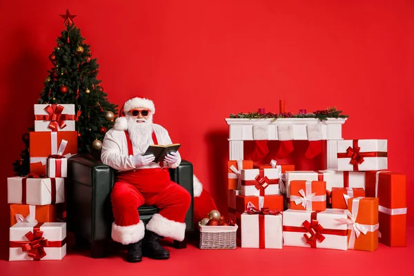 Portret van zijn hij aantrekkelijke funky vet overgewicht vrolijke Kerstman zitten in fauteuil lezen boek besteden feestelijke dag winter geïsoleerde helder levendig glans levendige rode kleur achtergrond — Stockfoto