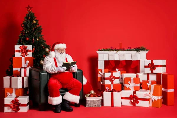 他那迷人的胖胖胖的形象，专注而快乐的圣诞老人坐在扶手椅上看书，花时间装饰客厅，明亮而生动的红色背景 — 图库照片