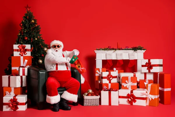 彼の魅力的な脂肪太りすぎファンキーな驚きの肖像サンタはアームチェアに座っている靴下の贈り物を詰めクリスマスに孤立した明るい鮮やかな輝き鮮やかな赤い色の背景 — ストック写真