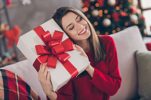 Pozitif kızın resmi Divan 'da büyük bir hediye kutusu tut. Evde kırmızı süveter giy ve Noel süsleri tak. — Stok fotoğraf